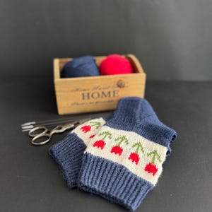 May include: A pair of blue knitted socks with a white band and red cherry designs. The socks are sitting on a black surface next to a wooden box with yarn and the word "HOME" printed on it.