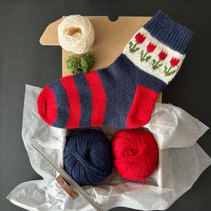Puede incluir: Un kit de tejido con un calcetín a rayas azules y rojas, dos ovillos de lana, uno blanco y uno rojo, y agujas de tejer. El calcetín tiene un puño blanco con un patrón de tulipanes rojos.