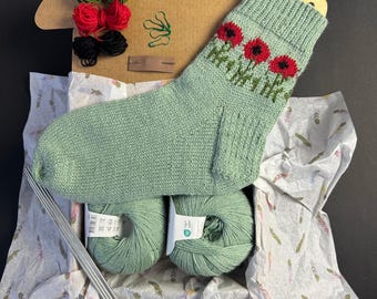 May include: A knitting kit containing a light green sock with red poppy detailing. The kit includes green yarn, knitting needles, and colourful yarn skeins. The items are arranged in a cardboard box lined with patterned tissue paper.