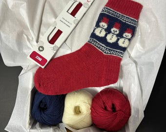 May include: A knitting kit for making a pair of red and blue snowman socks. The kit includes three balls of yarn in red, white, and blue, a pair of red double pointed knitting needles, and a red sock with a snowman pattern.