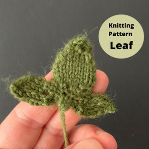 May include: A green knitted leaf with a pointed tip and two side leaves. The leaf is held in a hand and the yarn tail is visible.