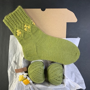 Könnte beinhalten: Eine grüne Stricksocke mit gelber Blumenstickerei, präsentiert in einer Box mit Garn und Nadeln. Die Socke ist hellgrün, mit geripptem Bündchen und detaillierter Blumenstickerei. Zwei Knäuel passendes grünes Garn und Stricknadeln sind enthalten.