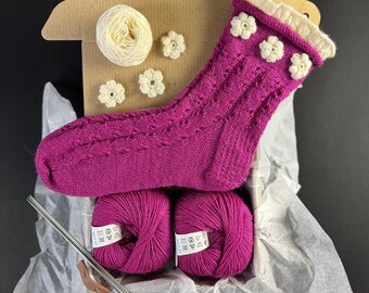 May include: A pink knitted sock with white crocheted flowers, two balls of pink yarn, and knitting needles in a cardboard box.