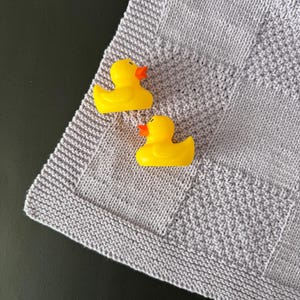 Puede incluir: Dos patitos de goma amarillos con picos naranjas están colocados sobre una manta de punto gris claro. La manta tiene un patrón texturizado y un borde sencillo. Los patitos están colocados cerca de la esquina de la manta.