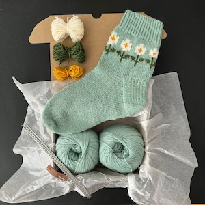 Puede incluir: Un kit de tejido para un par de calcetines verdes con un patrón de margaritas blancas. El kit incluye dos ovillos de lana verde, cuatro agujas de tejer y tres ovillos de lana blanca, verde y amarilla.