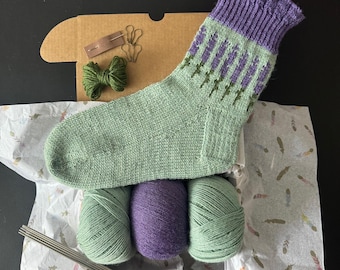 May include: A knitting kit with a light green and purple patterned sock. The kit includes green and purple yarn, knitting needles, a green yarn bow, and a small cardboard box with accessories. The sock is presented on patterned tissue paper, with a dark background.