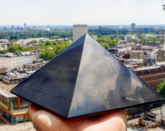 May include: A black obsidian pyramid crystal, with a smooth, polished surface, is held in a hand against a backdrop of a city skyline.