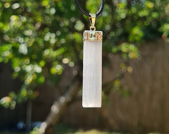 May include: A white selenite crystal pendant on a black cord necklace. The crystal is rectangular in shape and has a gold-toned metal setting.