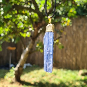 May include: A blue kyanite crystal pendant on a black cord necklace. The pendant is attached to a gold-colored bail.