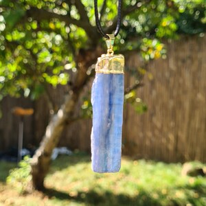 May include: A blue crystal pendant on a black cord necklace. The crystal is a long, rectangular shape with a gold-colored metal top.