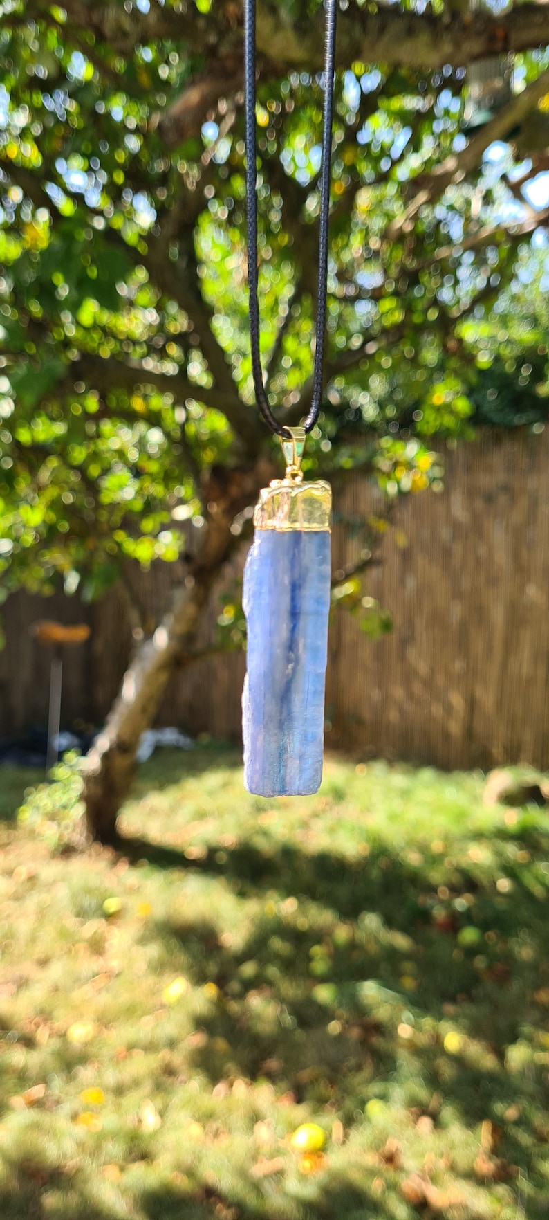 May include: A blue crystal pendant on a black cord necklace. The crystal is a long, rectangular shape with a gold-colored metal top.