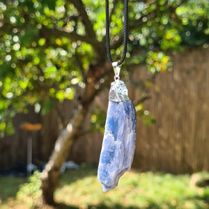 May include: A blue kyanite crystal pendant on a black cord necklace. The pendant is a raw, natural crystal with a silver bail.