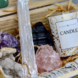 May include: A wooden box filled with crystals and a candle. The crystals include amethyst, selenite, rose quartz, and black tourmaline. The candle has a white label with the word "CANDLE" printed on it.