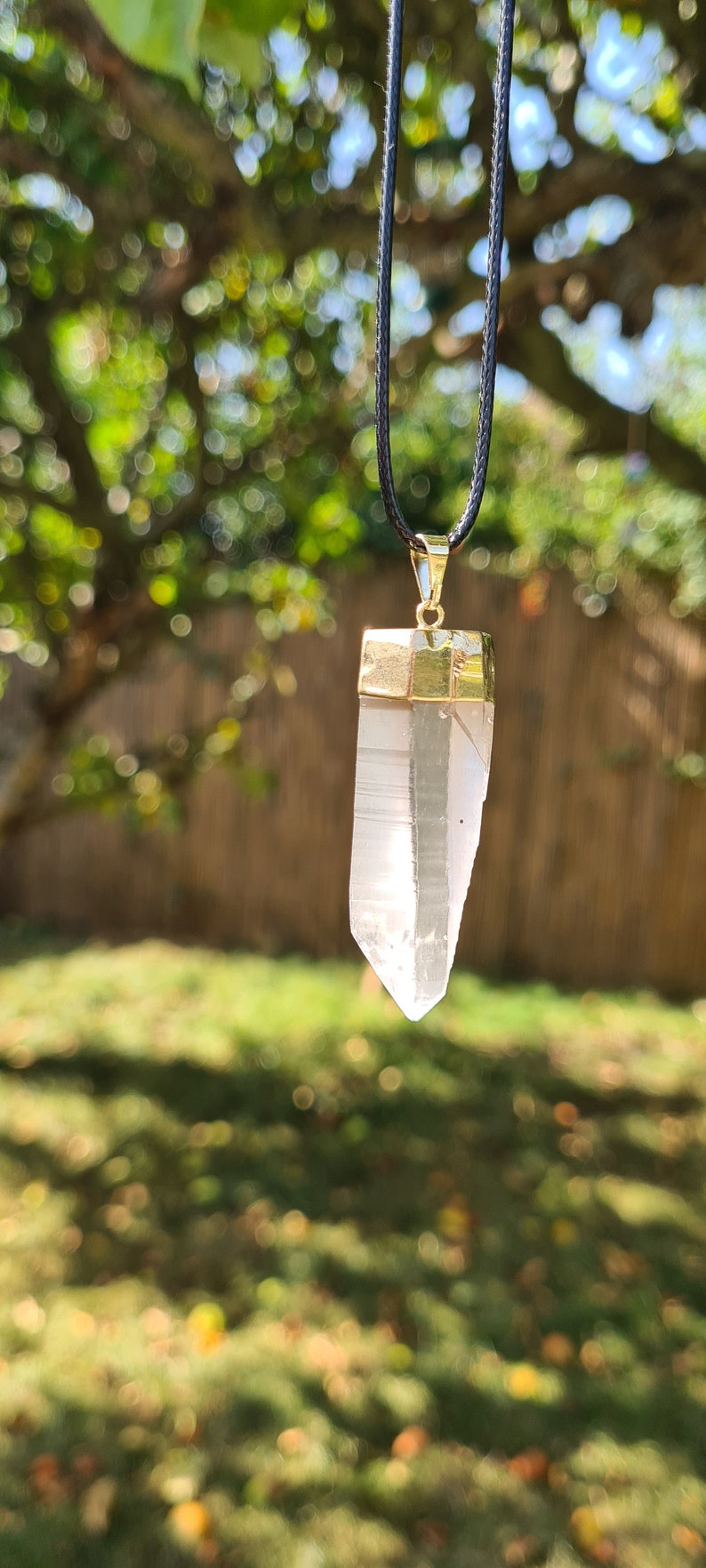 Puede incluir: Un colgante de cristal de cuarzo transparente con un engaste dorado cuelga de un cord&oacute;n negro. El colgante est&aacute; enfocado, mientras que el fondo est&aacute; desenfocado y muestra un &aacute;rea de hierba verde y un &aacute;rbol con hojas desenfocadas.