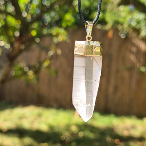 Puede incluir: Un colgante de cristal de cuarzo transparente con un engaste dorado cuelga de un cord&oacute;n negro. El colgante est&aacute; enfocado, mientras que el fondo est&aacute; desenfocado y muestra un &aacute;rea de hierba verde y un &aacute;rbol con hojas desenfocadas.