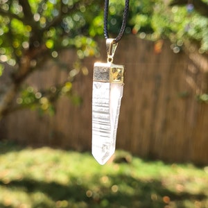 Puede incluir: Un colgante de cristal de cuarzo transparente en un collar de cord&oacute;n negro. El cristal est&aacute; facetado y tiene un engaste dorado.