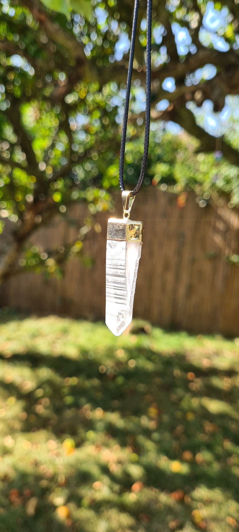 Puede incluir: Un colgante de cristal de cuarzo transparente en un collar de cord&oacute;n negro. El cristal est&aacute; facetado y tiene un engaste dorado.