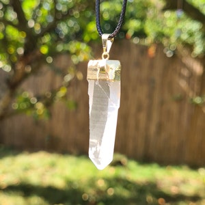 Puede incluir: Un colgante de cristal de cuarzo transparente en un collar de cord&oacute;n negro. El cristal est&aacute; engastado en un bisel de metal dorado.