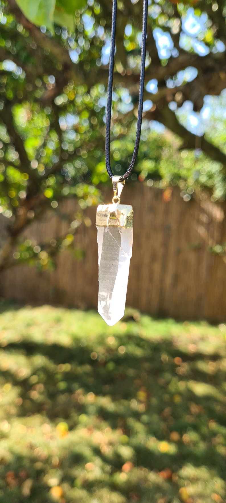 Puede incluir: Un colgante de cristal de cuarzo transparente en un collar de cord&oacute;n negro. El cristal est&aacute; engastado en un bisel de metal dorado.