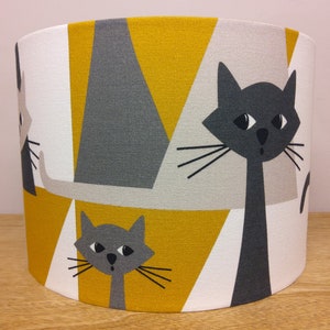 May include: Cylindrical lampshade with a repeating pattern of stylized black and grey cats on a geometric background. The background features triangles in white, mustard yellow, and grey. The lampshade is designed for a table lamp.