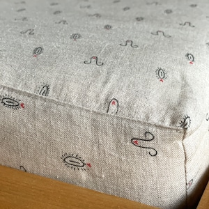 Könnte beinhalten: Nahaufnahme eines hellgrauen Leinenkissens mit einem schwarzen und roten Muster. Das Muster ist ein einfaches Design aus einem Kreis mit einer Linie durch ihn und einem kleinen Herzen oben. Das Kissen ist mit weißem Garn genäht und hat eine saubere, fertige Kante.