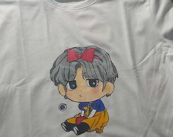 May include: White t-shirt with a hand-painted illustration of a cartoon character wearing a red bow and a yellow dress. The character is sitting down and holding a red apple.