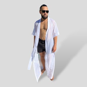 Op de afbeelding: Een witte, lichte badjas met een lang, vloeiend ontwerp. De badjas heeft korte mouwen en een strikceintuur. De persoon draagt er een zwarte zwembroek onder.