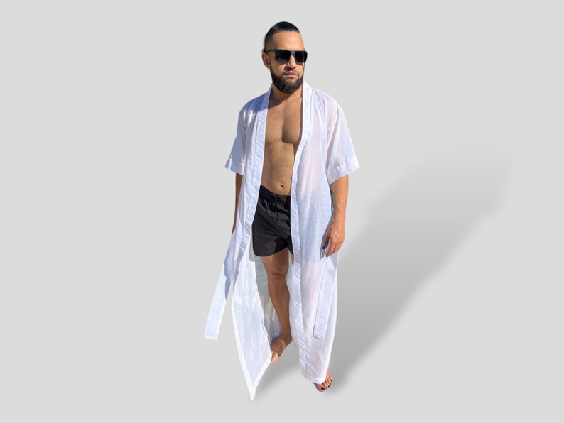 Op de afbeelding: Een witte, lichte badjas met een lang, vloeiend ontwerp. De badjas heeft korte mouwen en een strikceintuur. De persoon draagt er een zwarte zwembroek onder.