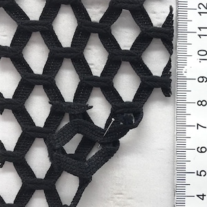 May include: Close-up of black netting with a honeycomb pattern. A ruler with centimetre measurements is visible along the right side. A pin is inserted into the netting. The netting is on a white background.