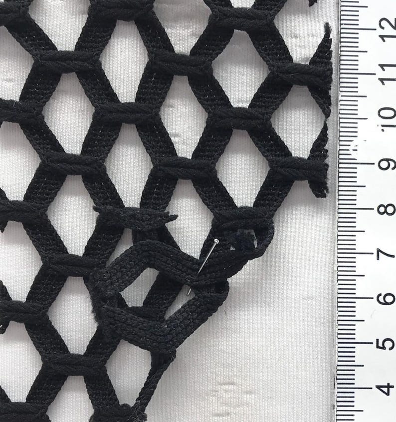 May include: Close-up of black netting with a honeycomb pattern. A ruler with centimetre measurements is visible along the right side. A pin is inserted into the netting. The netting is on a white background.