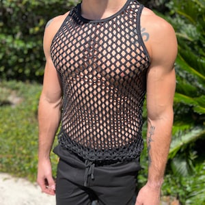 May include: Black fishnet vest and black shorts. The vest features a round neckline and a large open weave pattern. The shorts have a drawstring waist. The background is blurred with green foliage.
