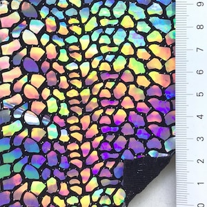 May include: A close-up of iridescent fabric with a black background and a pattern of small, irregular shapes. The fabric is textured and has a slight sheen. A ruler is visible in the background, with markings up to 9 inches.