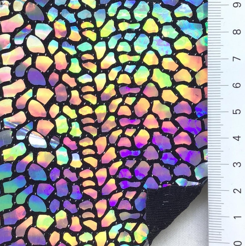 May include: A close-up of iridescent fabric with a black background and a pattern of small, irregular shapes. The fabric is textured and has a slight sheen. A ruler is visible in the background, with markings up to 9 inches.