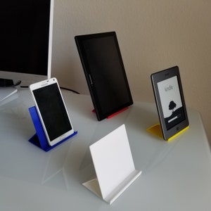 May include: Three different colored phone and tablet stands, made of plastic. The stands are blue, red, and yellow. The stands are holding a white smartphone, a black tablet, and a black e-reader. The e-reader has the word "kindle" on the screen.