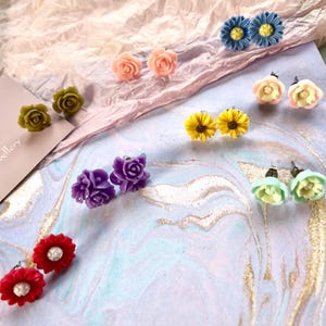 May include: A selection of floral stud earrings in a variety of colours. The earrings feature rose, sunflower, and daisy designs in pink, yellow, blue, green, purple, and red. The earrings are presented on a marbled surface.