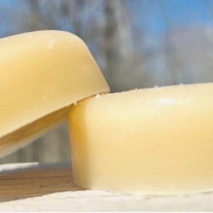 May include: Two pale yellow, oval-shaped bars of soap are displayed. One bar is leaning against the other, creating a slight angle. The soap has a smooth, slightly textured surface, and the background is blurred, suggesting an outdoor setting.