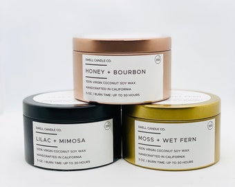 May include: Three scented candles in metallic tins. The top candle is copper-coloured, labelled "Honey + Bourbon." Below it is a black tin labelled "Lilac + Mimosa," and a gold tin with "Moss + Wet Fern." Each candle is 142 grams.