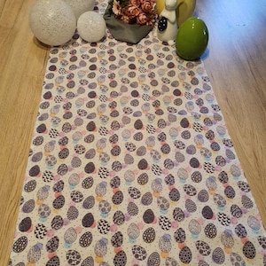 Può includere: Un runner da tavolo bianco con un motivo di uova di Pasqua colorate. Il runner è decorato con un piccolo bouquet di fiori rosa, una statuetta di coniglio bianco e tre sfere decorative bianche.