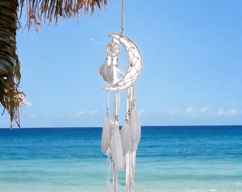 May include: White dreamcatcher with feathers hanging from a palm tree branch over a white sandy beach with blue ocean water in the background.