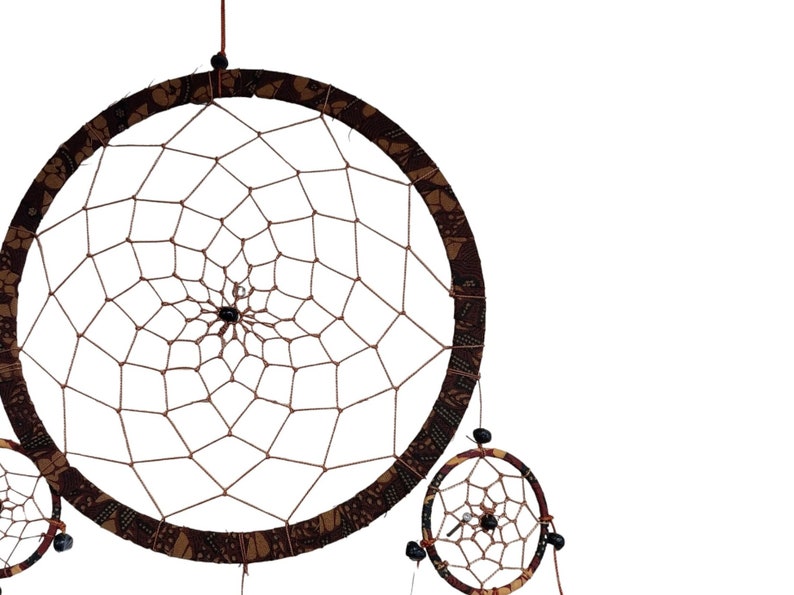 May include: A brown and tan dreamcatcher with a beaded fringe. The dreamcatcher has a circular frame with a web-like pattern and a smaller dreamcatcher hanging below it.