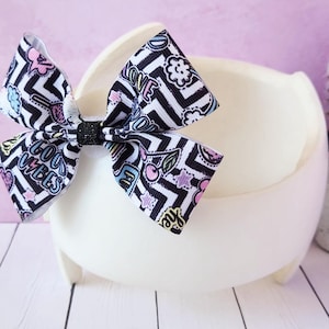 May include: A large, decorative hair bow with a black and white chevron pattern and colorful graphic accents. The bow features the words "Good Vibes" and "Love". The bow is set on a white, three-legged pedestal.