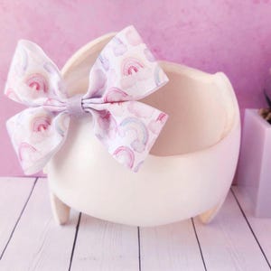 May include: A white ceramic bowl with a rainbow patterned bow on top. The bowl is sitting on a white wooden surface.