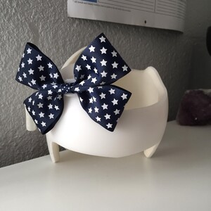 May include: A white, three-legged, rounded container with a navy blue bow featuring white stars. The bow is centered on the container, creating a patriotic or festive appearance. The container is on a white surface.