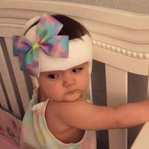 May include: A baby wearing a white padded headband with a colorful bow on top. The headband is designed to help shape the baby's head. The baby is wearing a tie-dye onesie and is looking to the side.