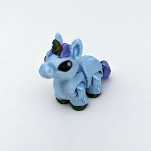 May include: A light blue 3D-printed unicorn figurine with a purple mane and tail, and a green horn. The unicorn has black eyes and green hooves. The figurine is posed on a white surface.