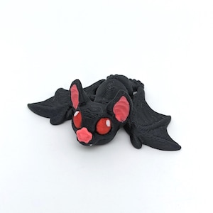 May include: A black bat figurine with red eyes, pink ears, and a pink nose. The bat has outstretched wings and is lying down. The figurine is made of a textured material and is set against a white background.