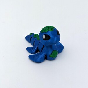 May include: A blue and green 3D-printed articulated caterpillar toy. The caterpillar has a blue body with green accents on its head and back. The eyes are black. The toy is on a white background.