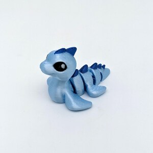 May include: A light blue, 3D-printed dragon figurine with a segmented body and large, black eyes. The dragon has a small head with pointed ears and a curved tail. The figurine is made of a shiny, textured material.