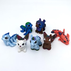 May include: A collection of colorful fantasy creature figurines. The assortment includes a blue dragon, a purple bird-like creature, a white cat-like figure, a blue unicorn, a brown winged creature, and a red dragon. Each figure is painted with vibrant colors and intricate details.