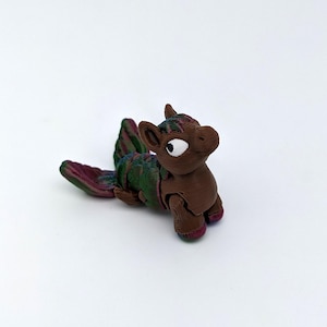 May include: A brown, stylized figurine of a mythical creature with a long tail and wings. The creature has a large eye and is lying down. The tail and wings are green, red, and purple. The figurine is on a white background.